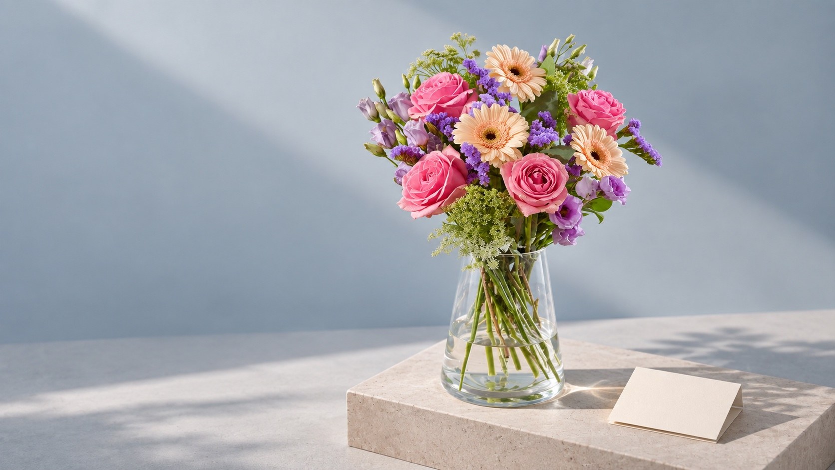Pink roses, peach gerberas and purple flowers in a clear vase beside a blank note card
