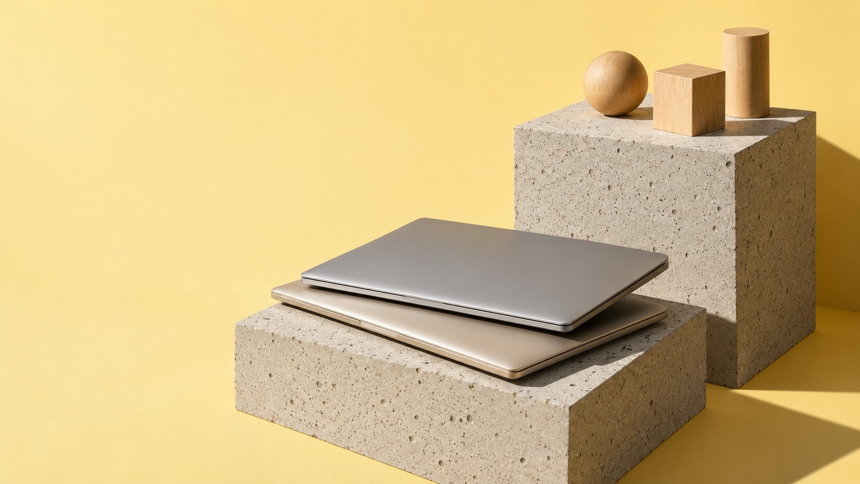 Two closed slim aluminium laptops stacked crosswise on a concrete block, beside a wooden sphere, cube and cylinder on a taller block, against a butter-yellow backdrop in hard afternoon light